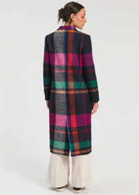 A woman shown from the back wearing a long plaid coat with a multicolor tartan pattern in navy, pink, green, orange, and white, paired with cream wide-leg pants, against a plain light gray studio background.