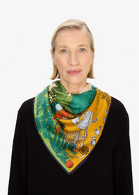 A woman with short blonde hair wearing a black sweater and a vibrant, patterned silk scarf. The scarf features a mix of green, yellow, orange, and blue botanical and animal illustrations, styled against a clean white background.