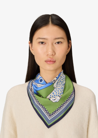 A model wearing a green silk scarf with a blue and white spotted fish pattern, styled around the neck over a cream-colored knit sweater against a plain light gray background.