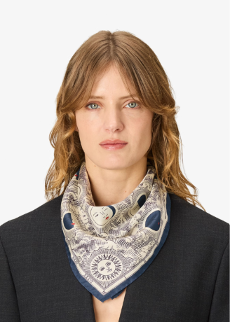 A woman with blonde hair wearing a patterned silk scarf with a blue border and a dark gray blazer, photographed against a plain white background.