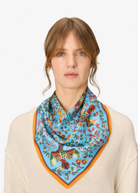 A woman wearing a light blue silk scarf with a colorful bird and floral print and a yellow-orange border, paired with a cream-colored knit sweater against a plain white background.