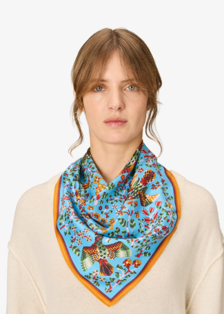 A woman wearing a light blue silk scarf with a colorful bird and floral print and a yellow-orange border, paired with a cream-colored knit sweater against a plain white background.