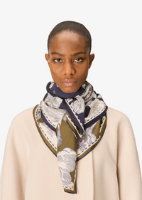 A model wearing a cream silk scarf with a navy blue border and olive green patterned details, styled with a beige sweater against a light gray background.