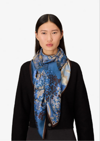A model wearing a black cardigan and a blue patterned silk scarf with floral and paisley designs, set against a plain light gray background.