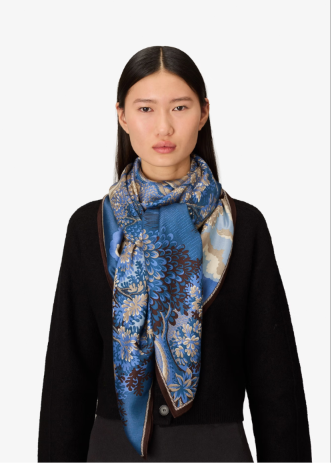 A model wearing a black cardigan and a blue patterned silk scarf with floral and paisley designs, set against a plain light gray background.