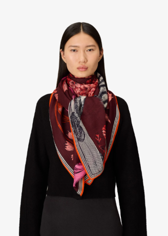 A model wearing a dark burgundy silk scarf with a floral print, striped pattern, and orange border, styled over a black top against a light gray background.