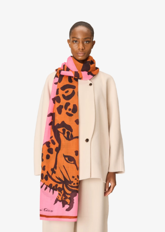 A model wearing a cream-colored coat and trousers, styled with a vibrant pink and orange scarf featuring a leopard print and a large tiger face design. The brand name 'Sandro' is visible in script near the bottom corner of the scarf.