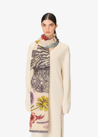A model wearing a cream-colored knit cardigan and a long cream scarf with a tiger and floral print, set against a plain light gray background.