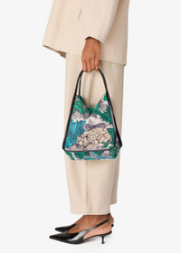 A person wearing beige pants and a matching blazer holds a green and teal patterned shoulder bag with black handles. The person is also wearing black slingback heels against a plain light gray studio background.