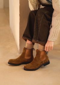 A person seated indoors wearing brown suede Chelsea-style ankle boots with elastic side panels, paired with dark brown corduroy pants and beige socks.
