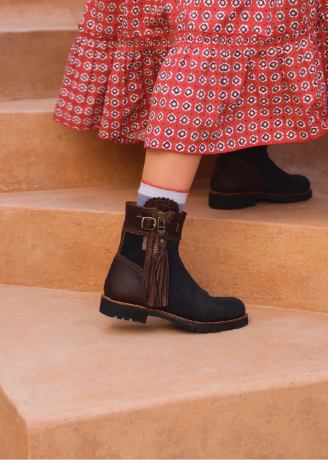 A close-up of a person wearing dark navy and brown leather ankle boots with a side zipper and tassel detail, standing on tan stone steps. The person is also wearing a red patterned skirt and a light blue sock.