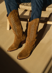 A pair of tan suede cowboy boots with silver embroidered leaf patterns, worn with blue jeans while sitting on a woven chair in warm sunlight.
