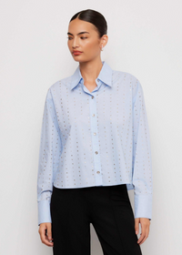 A woman modeling a light blue long-sleeve button-up shirt with a subtle repeating geometric pattern, paired with black trousers, against a plain white studio background.