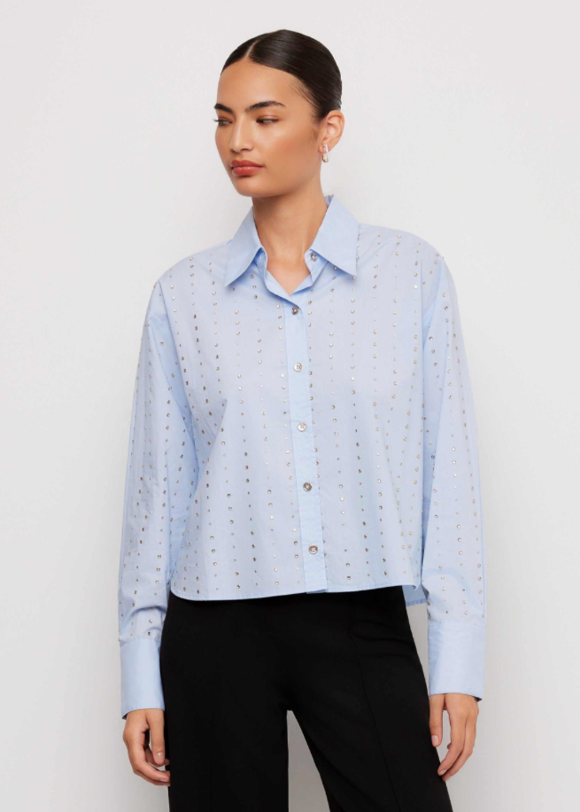 A woman modeling a light blue long-sleeve button-up shirt with a subtle repeating geometric pattern, paired with black trousers, against a plain white studio background.