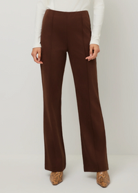 A model wearing a pair of high-waisted, wide-leg brown trousers, styled with a white ribbed long-sleeve top and brown patterned pointed-toe heels, shot against a neutral light gray studio background.