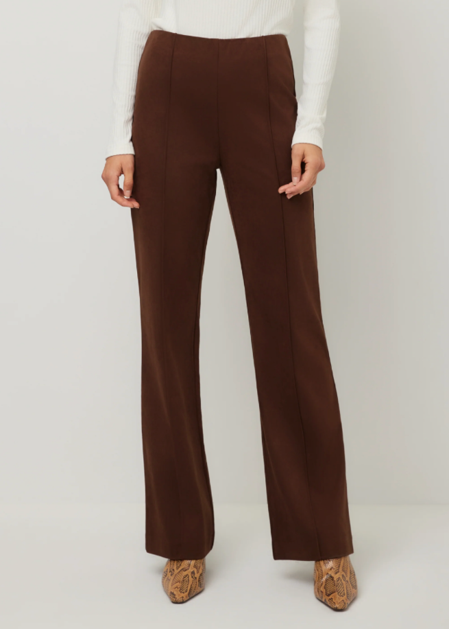 A model wearing a pair of high-waisted, wide-leg brown trousers, styled with a white ribbed long-sleeve top and brown patterned pointed-toe heels, shot against a neutral light gray studio background.
