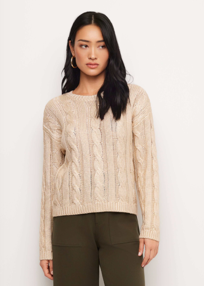 A woman modeling a cream cable-knit crewneck sweater with olive green pants, standing against a plain white studio background.