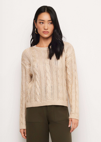 A woman modeling a cream cable-knit crewneck sweater with olive green pants, standing against a plain white studio background.