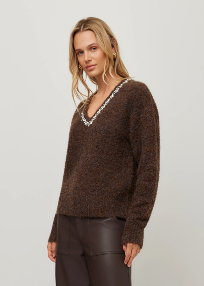 A woman modeling a brown fuzzy knit sweater with a white beaded V-neck trim, paired with dark brown pants, against a plain light gray background.
