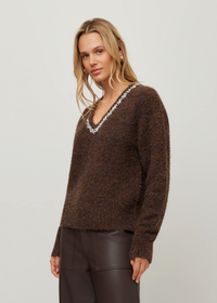 A woman modeling a brown fuzzy knit sweater with a white beaded V-neck trim, paired with dark brown pants, against a plain light gray background.