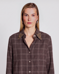 A woman wearing a brown plaid button-up shirt with gold-tone stud embellishments on the collar, photographed against a light gray studio background.