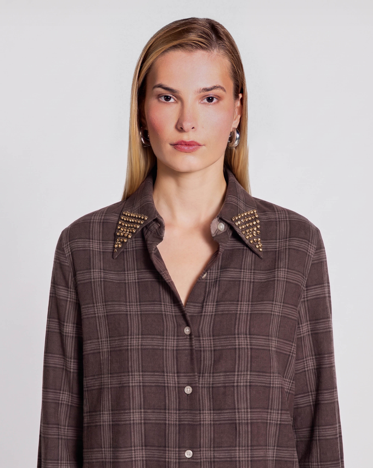 A woman wearing a brown plaid button-up shirt with gold-tone stud embellishments on the collar, photographed against a light gray studio background.