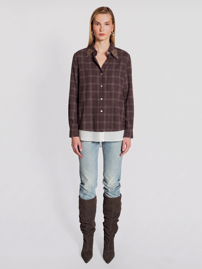 A model stands against a light gray studio background wearing a dark brown and black plaid button-up shirt with patterned shoulder details, light-wash blue jeans, and dark brown over-the-knee boots.