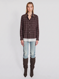 A model stands against a light gray studio background wearing a dark brown and black plaid button-up shirt with patterned shoulder details, light-wash blue jeans, and dark brown over-the-knee boots.