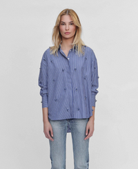 A woman modeling a blue and white vertically striped button-up shirt with small embroidered details, paired with light-wash jeans, against a plain light gray background.