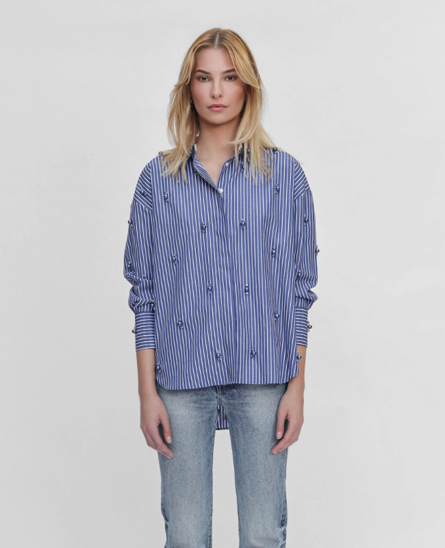 A woman modeling a blue and white vertically striped button-up shirt with small embroidered details, paired with light-wash jeans, against a plain light gray background.