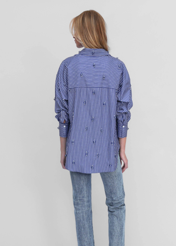 A woman viewed from the back wearing a blue and white striped button-up shirt with embroidered details, paired with light-wash blue jeans, against a plain light gray studio background.