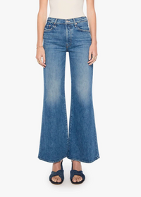 A model wearing a pair of medium-wash blue denim flare jeans, paired with a white top and dark blue sandals, shot against a plain white studio background.