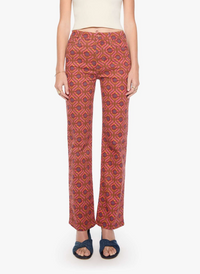 A model is shown from the waist down wearing a pair of straight-leg pants with a red, orange, and blue geometric pattern, paired with a cream top and dark blue slide sandals, against a plain white background.