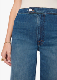Close-up of a person wearing medium blue denim jeans with a visible front pocket, button waistband, and hand resting near the leg against a plain light background.