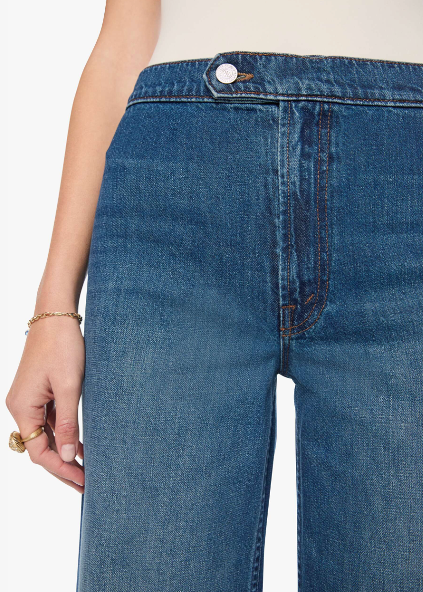 Close-up of a person wearing medium blue denim jeans with a visible front pocket, button waistband, and hand resting near the leg against a plain light background.