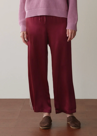 A person wearing burgundy wide-leg satin pants with white side stripes, a pink ribbed sweater, and brown slip-on shoes, standing against a plain light wall and wood floor.
