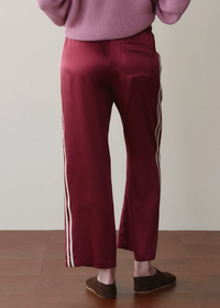 A person shown from the back wearing burgundy satin pants with white side stripes, a pink ribbed sweater, and brown slip-on shoes against a plain beige wall and wood floor.