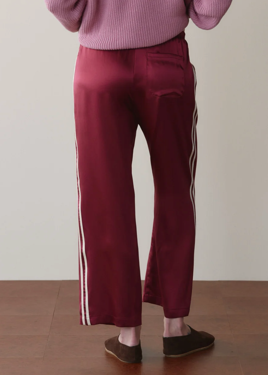 A person shown from the back wearing burgundy satin pants with white side stripes, a pink ribbed sweater, and brown slip-on shoes against a plain beige wall and wood floor.