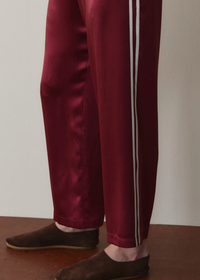A person wearing burgundy satin track pants with white side stripes and dark brown suede slip-on shoes, standing on a wooden floor against a plain light wall.
