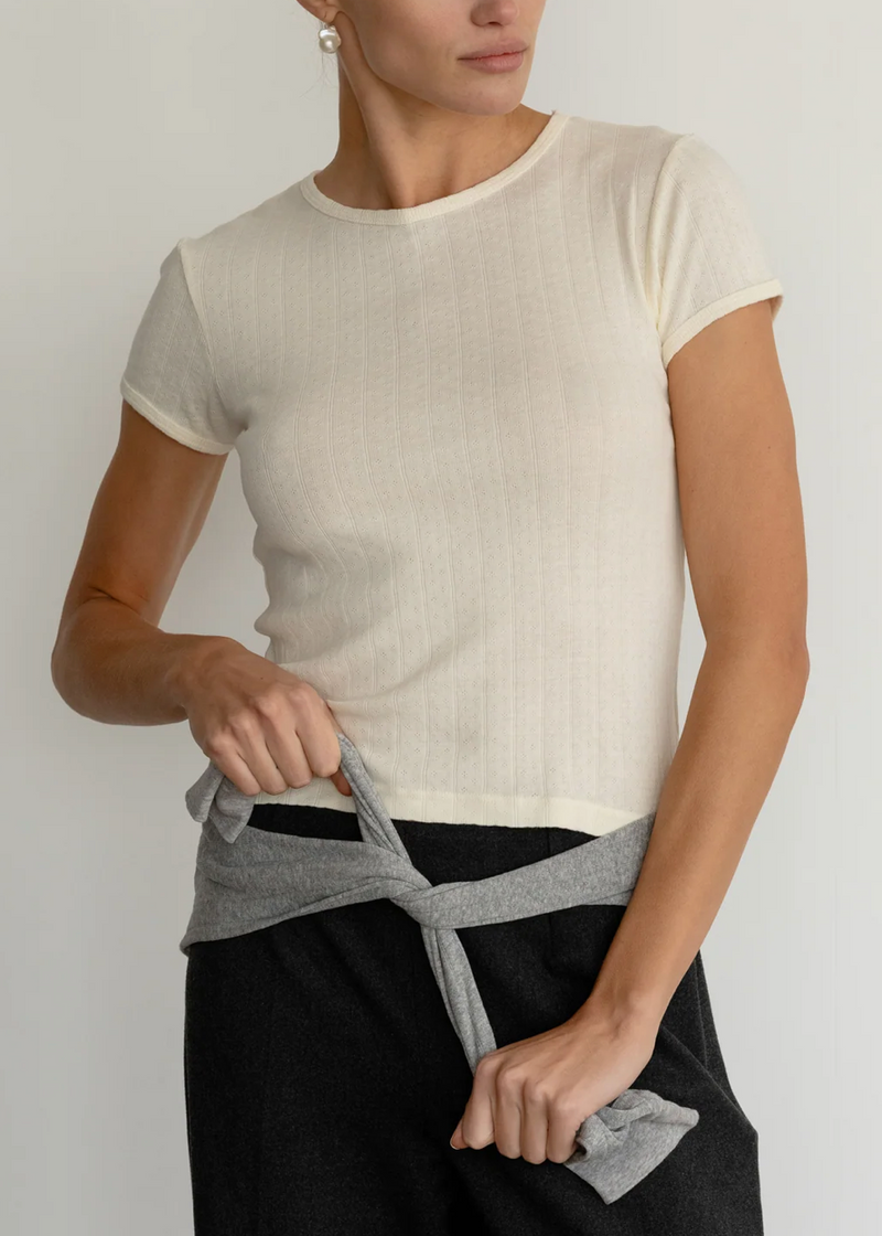 A woman wearing a fitted short-sleeve cream ribbed knit top and black pants, tying a gray knit garment around her waist against a plain light background.