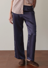 A person wearing dark navy satin pants with a subtle sheen, paired with a beige and white gingham shirt and brown suede shoes, standing against a neutral beige wall and wooden floor.