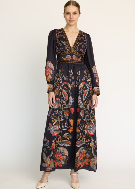 A woman modeling a long-sleeved maxi dress with a dark navy base and an intricate multicolor floral and botanical print, styled with beige pointed-toe heels against a plain white studio background.