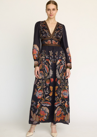 A woman modeling a long-sleeved maxi dress with a dark navy base and an intricate multicolor floral and botanical print, styled with beige pointed-toe heels against a plain white studio background.