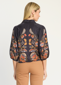 A woman shown from the back wearing a dark navy floral blouse with puff sleeves and tan pants, photographed against a plain light background.