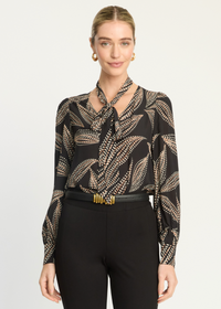 A woman modeling a black long-sleeve blouse with a beige and gold abstract leaf pattern, paired with black trousers and a gold belt, against a light gray studio background.