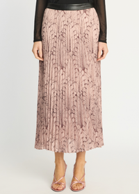 A model wearing a pleated midi skirt in dusty pink with a dark brown abstract leaf pattern, styled with a black long-sleeve top and pink strappy heels against a light gray studio background.