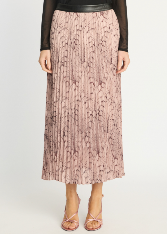 A model wearing a pleated midi skirt in dusty pink with a dark brown abstract leaf pattern, styled with a black long-sleeve top and pink strappy heels against a light gray studio background.