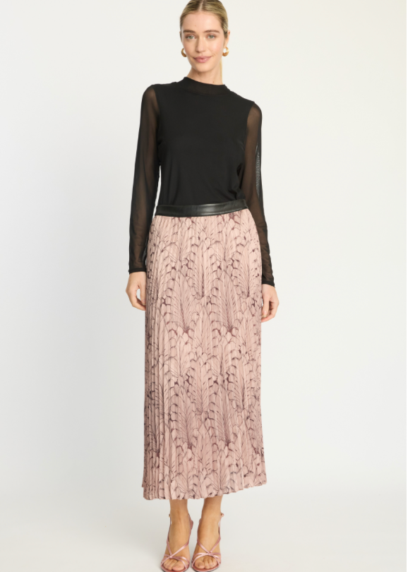 A model stands against a light gray studio background wearing a black sheer long-sleeve top with a black faux-leather waistband and a pleated dusty pink maxi skirt with a dark abstract leaf pattern, paired with metallic pink heeled sandals.