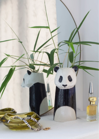 A decorative panda-shaped vase with a glossy black and white finish, shown with bamboo stems in a styled interior setting. A mirror behind it reflects the back of the vase, and small accessories including a glass dish, bracelet, and perfume bottle sit nearby on a white surface.