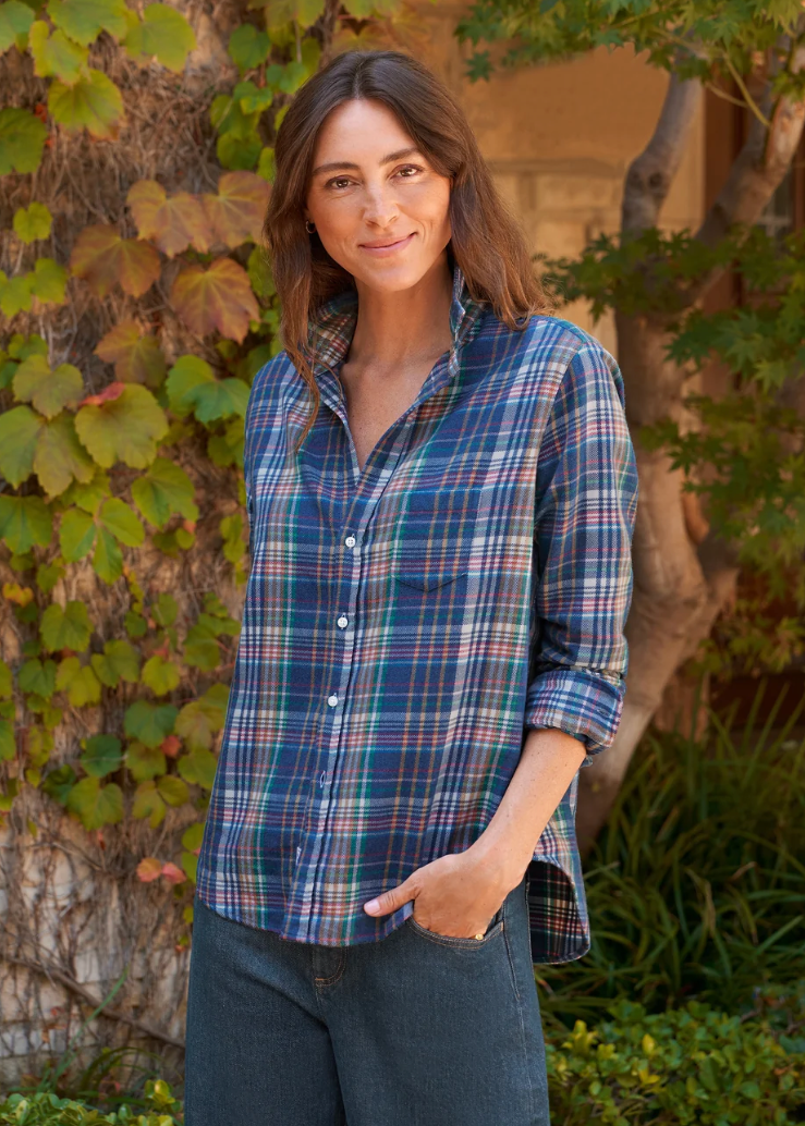 A woman wearing a blue and green plaid button-down shirt with dark jeans, standing outdoors in front of a tree with green and yellow leaves.